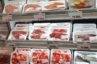 Balcão refrigerado de supermercado com bandejas de carne fresca e congelada embaladas, rótulos e preços visíveis.